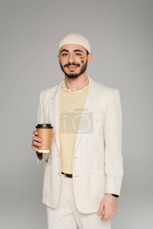 Fashionable gay man with lgbt flag on cheek holding coffee to go isolated on grey  