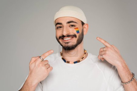 Smiling homosexual man in hat pointing at lgbt flag on cheek isolated on grey  