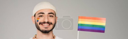 Cheerful homosexual man in hat looking at camera near lgbt flag isolated on grey, banner 