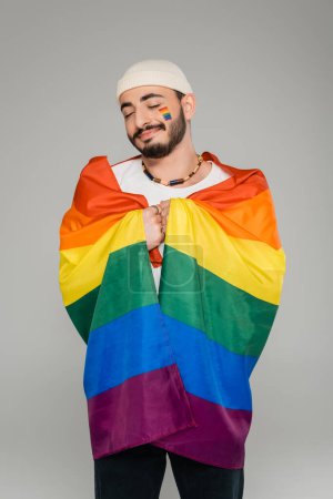 Fröhlicher schwuler Mann mit lgbt Flagge und geschlossenen Augen isoliert auf grau  