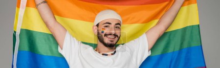Fröhlicher schwuler Mann mit Hut, der die LGBT-Fahne hält und isoliert auf graues Banner in die Kamera blickt 