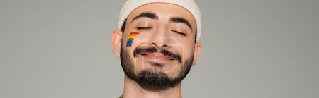 Smiling homosexual man in hat with lgbt flag on cheek isolated on grey, banner 