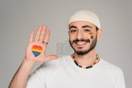 Lächelnder homosexueller Mann mit Herzzeichen von der lgbt-Fahne an der Hand isoliert auf grau  