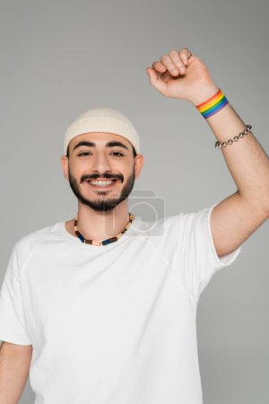 Cheerful gay man showing lgbt bracelet on hand isolated on grey  