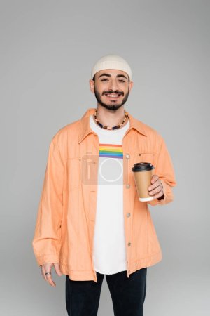 Smiling homosexual man with lgbt flag on t-shirt holding coffee to go isolated on grey  