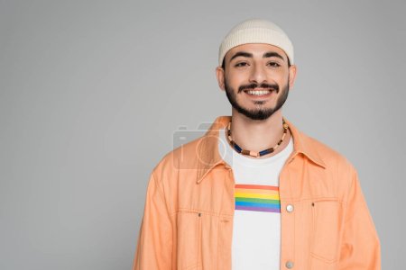 Lächelnder und stilvoller schwuler Mann mit lgbt-Flagge auf T-Shirt, der isoliert auf grau in die Kamera blickt  