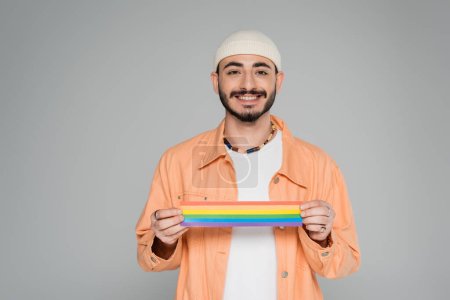 Fröhlicher homosexueller Mann mit lgbt-Fahne und Blick in die Kamera isoliert auf grau  