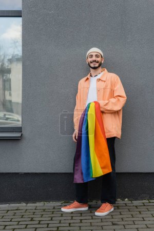 Lächelnder schwuler Mann mit lgbt-Fahne in der Nähe eines Gebäudes im Freien 
