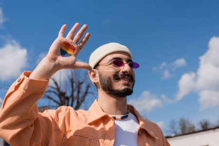 Cheerful gay man in sunglasses with lgbt flag in heart shape on hand standing outdoors 