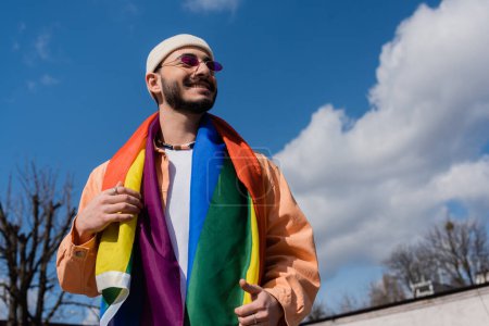 Vista de bajo ángulo del hombre homosexual sonriente en gafas de sol con bandera lgbt en la calle urbana, Día internacional contra la homofobia