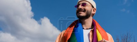 Foto de Vista de ángulo bajo del hombre gay despreocupado en gafas de sol con bandera lgbt de pie en la calle, pancarta - Imagen libre de derechos