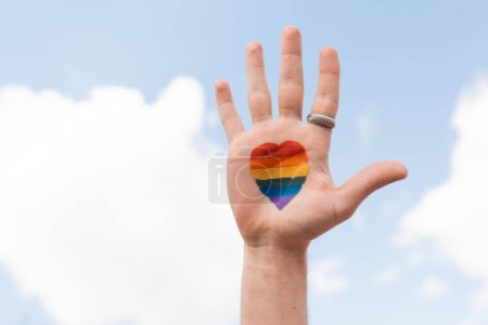 Téléchargez les photos : Cropped view of gay man with lgbt flag from heart sign and sky at background, Journée internationale contre l'homophobie - en image libre de droit