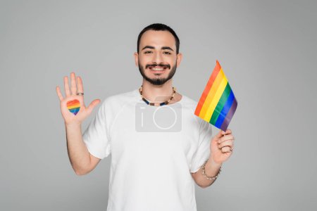 Smiling gay man with lgbt flag and heart on hand looking at camera isolated on grey, International day against homophobia
