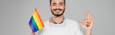Hombre homosexual alegre con bandera lgbt y corazón en la mano aislado en gris, bandera 