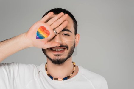 Lächelnder schwuler Mann bedeckt Augen mit lgbt-Fahne an der Hand isoliert auf grau, Internationaler Tag gegen Homophobie