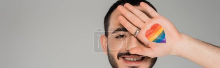 Photo for Cheerful homosexual man covering face with lgbt flag on hand isolated on grey, banner - Royalty Free Image