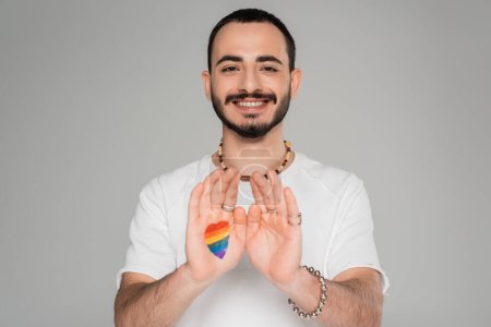 Young smiling gay man with lgbt flag on hand looking at camera isolated on grey, International day against homophobia