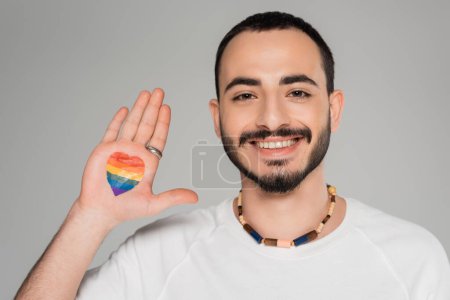 Joyeux jeune homme homosexuel avec drapeau lgbt à portée de main regardant la caméra isolée sur gris  