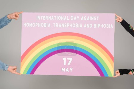 Téléchargez les photos : Vue recadrée de personnes homosexuelles portant une pancarte portant le lettrage "Journée internationale contre l'homophobie, la transphobie et la biphobie" isolé sur du gris - en image libre de droit