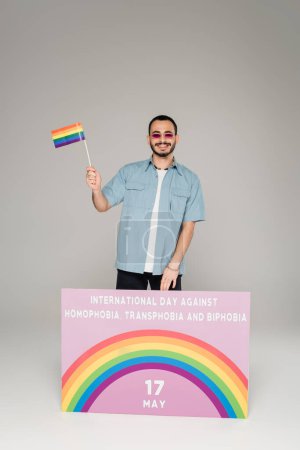 Fröhlicher schwuler Mann mit lgbt-Fahne neben Plakat mit Schriftzug gegen Homophobie, Transphobie und Biphobie auf grauem Hintergrund 