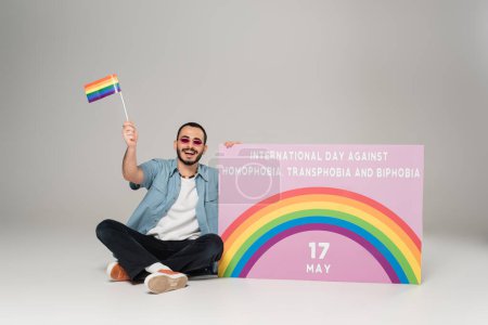 Hombre gay positivo sosteniendo bandera lgbt cerca de pancarta con el Día Internacional contra la Homofobia, Transfobia y Bifobia escrito en gris 