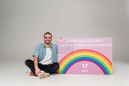 Fröhlicher Schwuler neben Plakat mit Schriftzug gegen Homophobie, Transphobie und Biphobie auf grau 