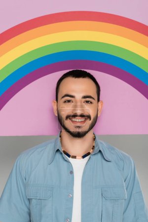 Foto de Retrato de hombre gay sonriente mirando a la cámara cerca de pancarta con bandera lgbt aislado en gris - Imagen libre de derechos