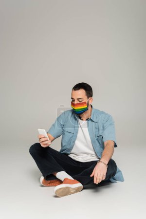 Homosexueller Mann in medizinischer Maske mit lgbt-Flaggenfarben mit Smartphone auf grauem Hintergrund 