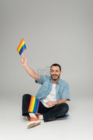 Smiling gay man holding lgbt flags while sitting on grey background 