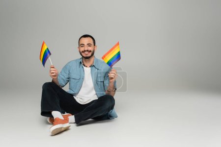 Alegre hombre homosexual sosteniendo banderas lgbt y mirando a la cámara mientras está sentado sobre fondo gris 