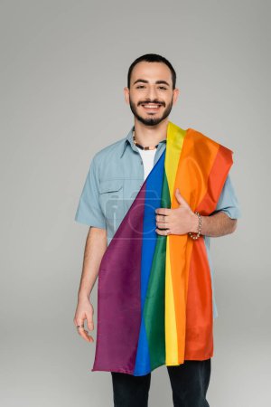Young bearded gay man with lgbt flag smiling at camera isolated on grey, international homophobia day  