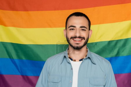 alegre gay hombre mirando a cámara cerca lgbt bandera en fondo, internacional día contra homofobia