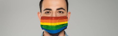 Young brunette gay man in medical mask with lgbt flag looking at camera isolated on grey, banner 