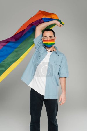 Young homosexual man in medical mask holding lgbt flag isolated on grey, international homophobia day  