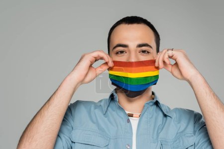 Hombre homosexual joven en máscara médica en colores de bandera lgbt aislado en gris  