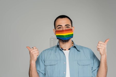 Hombre homosexual morena en máscara médica en colores lgbt mostrando como gesto aislado en gris, Día Internacional contra la Homofobia