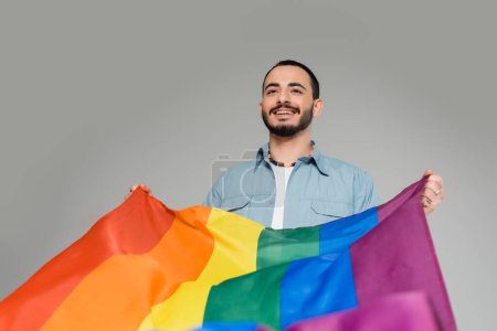 Cheerful young gay man holding lgbt flag isolated on grey, International Day Against Homophobia