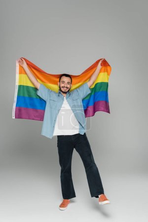 Full length of positive homosexual man holding lgbt flag on grey background 