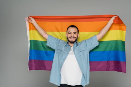 Joven homosexual sosteniendo bandera lgbt y mirando a la cámara aislada en gris, Día Internacional contra la Homofobia