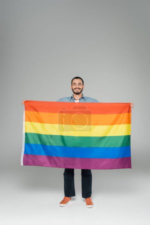 Pleine longueur de jeune homme gay tenant drapeau lgbt et souriant à la caméra sur fond gris, Journée internationale contre l'homophobie