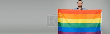 Morena gay hombre holding lgbt bandera y mirando a cámara aislado en gris, bandera 