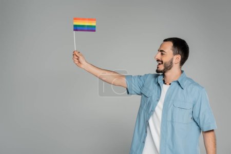 Téléchargez les photos : Vue latérale de l'homme homosexuel joyeux tenant drapeau lgbt et regardant loin isolé sur gris - en image libre de droit
