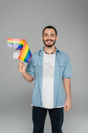 Sonriente hombre homosexual sosteniendo banderas lgbt y mirando a la cámara aislada en gris  