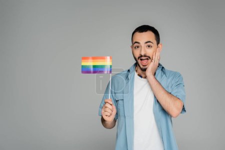 Erstaunter homosexueller Mann berührt Gesicht und hält lgbt Flagge isoliert auf grau, Internationaler Tag gegen Homophobie 
