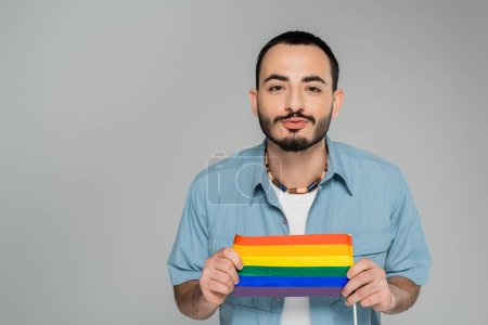 Morena gay hombre puchando labios y sosteniendo lgbt bandera aislado en gris, Día Internacional contra la Homofobia 