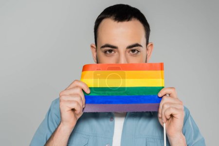 Photo for Young brunette gay man covering face with lgbt flag isolated on grey, International Day Against Homophobia - Royalty Free Image