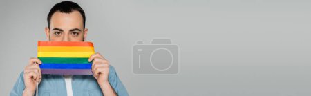 Téléchargez les photos : Brunette homosexuel homme couvrant visage avec drapeau lgbt isolé sur gris, bannière - en image libre de droit