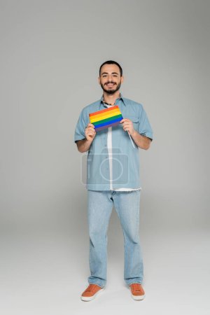 Full length of smiling homosexual man in denim shirt holding lgbt flag on grey background 