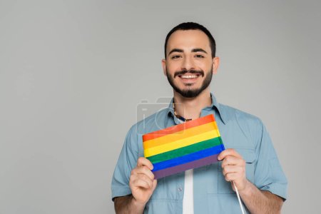 Portrait of cheerful gay man in shirt holding lgbt flag isolated on grey  
