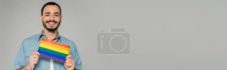 Téléchargez les photos : Homme homosexuel barbu en chemise tenant le drapeau lgbt et souriant isolé sur gris, bannière - en image libre de droit